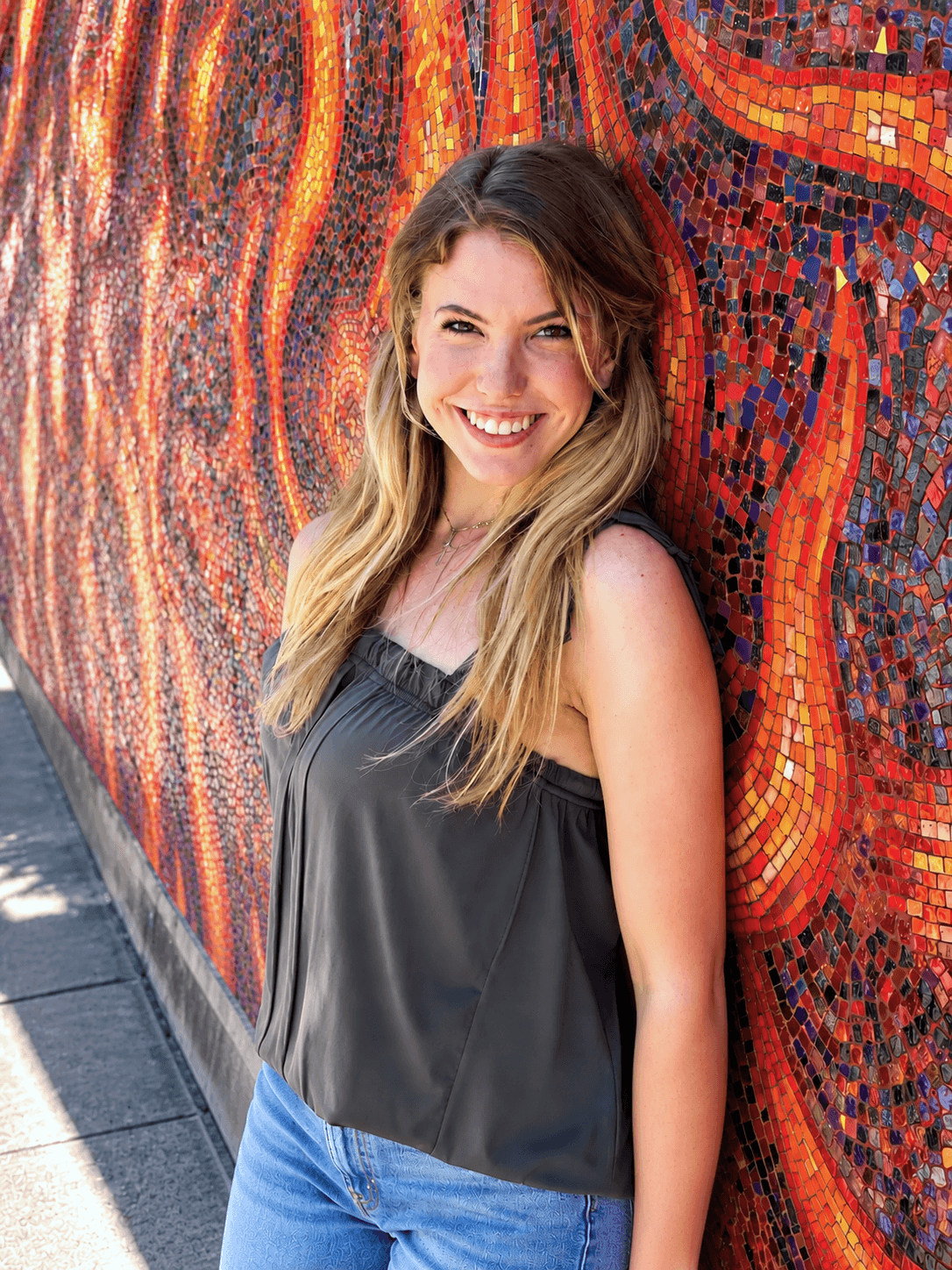 Ally Nowakowski smiling in front of a vibrant red-and-orange mosaic mural.